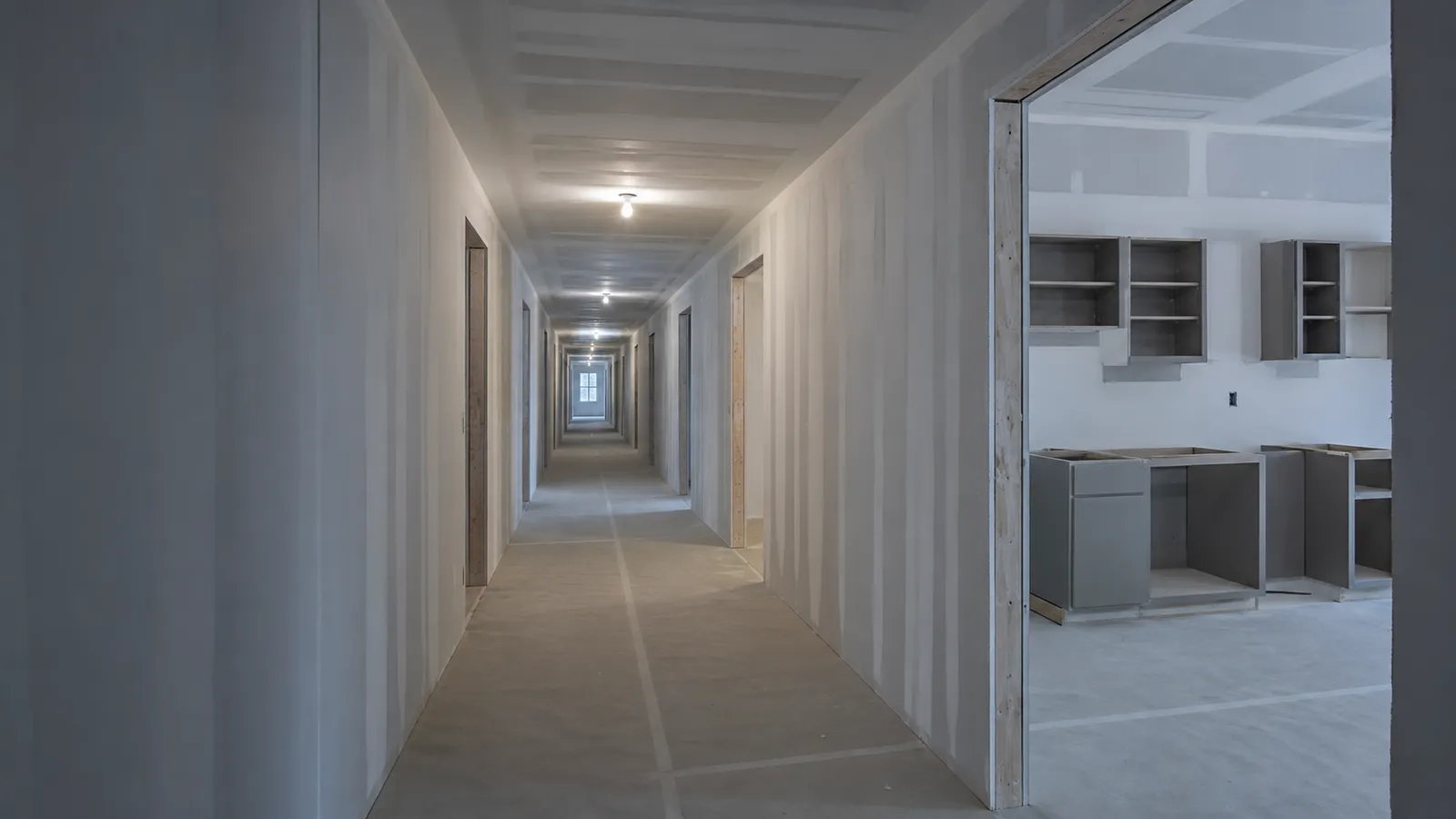 Corridor of a multifamily building during fit-out, with identical unit doorways and cabinet boxes set in an unfinished kitchen